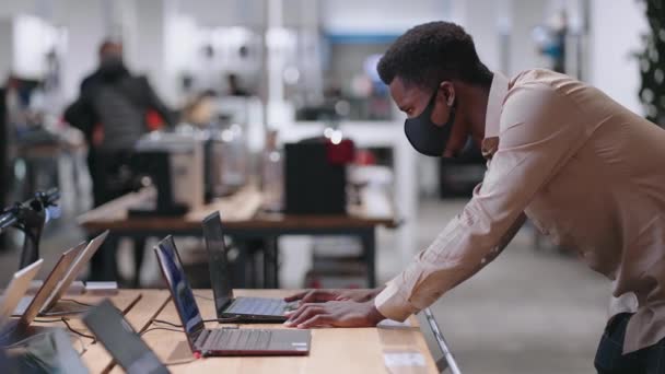 Negro chico con mascarilla está eligiendo el ordenador portátil moderno para el trabajo en la tienda de electrónica, prueba de muestra de exposición — Vídeo de stock