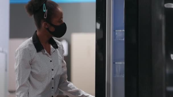 Afro-americká hospodyňka s obličejovou maskou si vybírá ledničku v obchodě s domácími spotřebiči, prohlížení exponátu uvnitř, střední portrét — Stock video