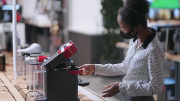 Женщина покупатель выбирает автоматический кофеварка в магазине бытовой техники, афро-американская леди с маской для лица просматривает образец выставки — стоковое видео