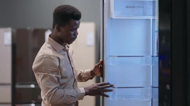 Erkek Afro-Amerikalı ziyaretçi ev aletleri mağazasından yeni buzdolabı seçiyor. Ticaret salonundaki sergi örneklerini inceliyor. — Stok video