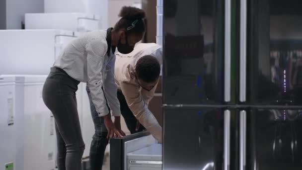 Negro hombre y mujer están de compras en la tienda de hardware, la elección de nuevo refrigerador en la tienda de electrodomésticos — Vídeos de Stock