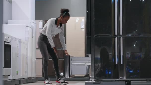 Afrikaans-Amerikaanse dame kiest koelkast in huis apparaat winkel, het bekijken van vriezer van de tentoonstelling monster binnen, full-length portret — Stockvideo