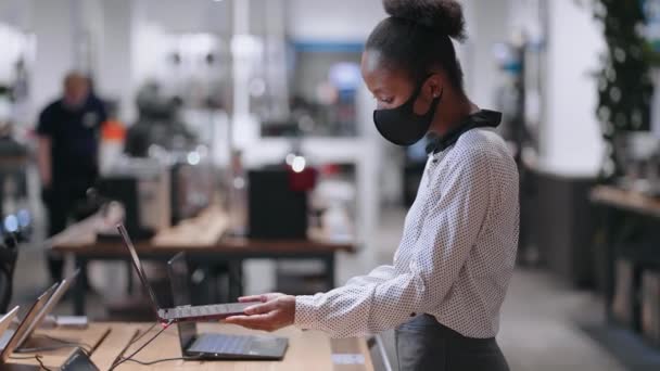 Giovane donna afro-americana con maschera facciale sta scegliendo il computer portatile moderno nel negozio di elettronica, visualizzazione campione mostra — Video Stock