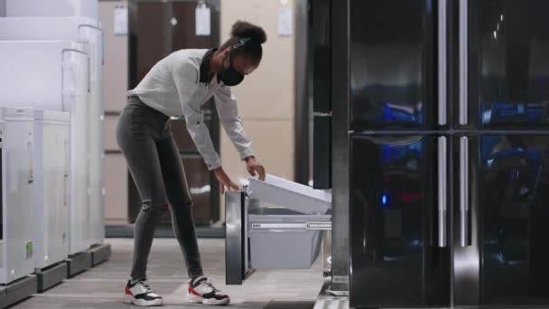 Jonge zwarte vrouw met gezichtsmasker is het openen van vriezer van de koelkast in huis apparaat winkel, het bekijken van de tentoonstelling monster binnen, full-length portret — Stockvideo