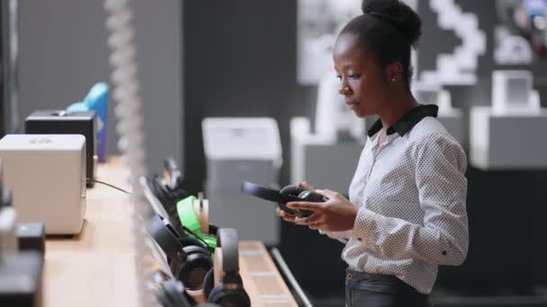 年轻的非洲裔美国女性将耳机放在家用电器商店的交易大厅里，在购买前先试用一下新产品 — 图库视频影像