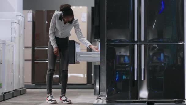 Genç siyahi kadın, ev aletleri mağazasında yeni bir buzdolabı modeli görüyor. İçerideki sergi örneğine bakıyor. Kadın alışverişçinin tam boy portresi. — Stok video