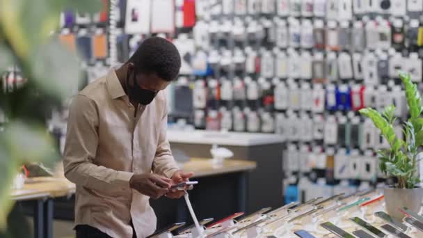 Afro-americano cara está comprando durante pandemia de covid-19 na loja de equipamentos digitais, escolhendo smartphone e pensando em comprar — Vídeo de Stock