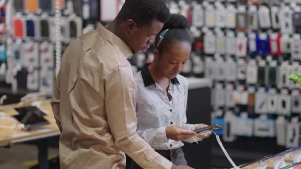 Afro-amerikaanse man helpt zijn vriendin om mobiele telefoon te kiezen, pair is winkelen in digitale apparatuur winkel — Stockvideo