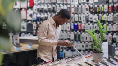 Afro-Amerikalı adam dijital ekipman mağazasında farklı akıllı telefon modellerini test ediyor. Cihazı seçiyor ve satın almayı düşünüyor.