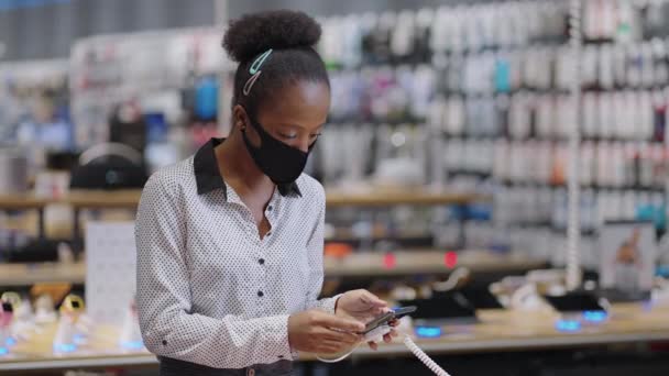 Elektronik mağazasında kadın alışverişçinin portresi covid-19 salgını sırasında, yüz maskeli siyah kadın akıllı telefonu seçiyor. — Stok video