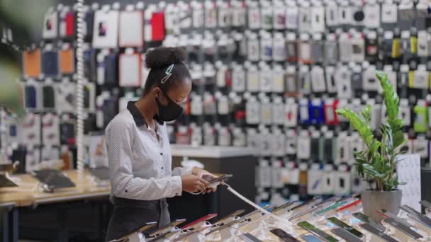 Afro-amerikan kadın alışverişçi cep telefonunu tercih ediyor. Elektronik dükkanında, covid-19 salgını sırasında, yüz maskeli siyah kadın. — Stok video