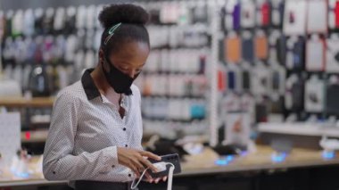Yüz maskeli Afro-Amerikalı kadın dijital ekipman mağazasında modern akıllı telefonu seçiyor.