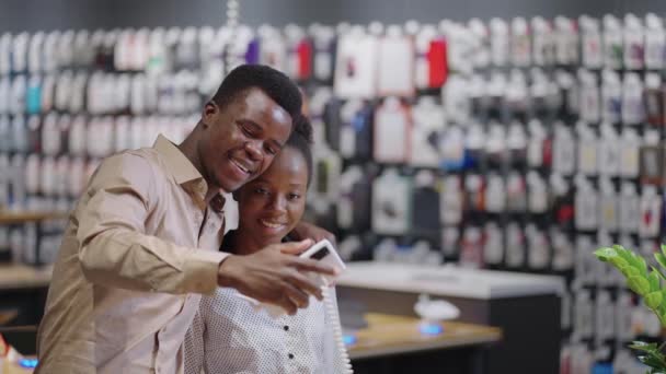 Happy afro-american couple in digital equipment store, shoppers are checking camera of exhibition sample of smartphone, taking selfie together together — Stock Video