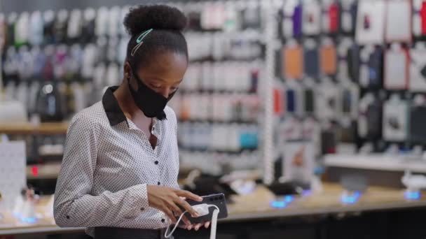 Afro-american woman with face mask is choosing modern smartphone in digital equipment store, viewing new models in trading hall before buying — Stock Video