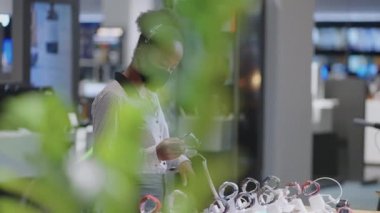 Yüz maskeli kadın elektronik mağazasından akıllı kaset alıyor. Gösteri vitrininde yeni modelleri inceliyor.