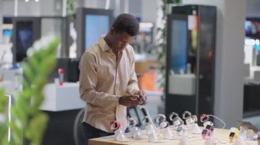 Afro-Amerikalı adam elektronik mağazasında akıllı saat seçiyor. Vitrindeki sergi örneklerine bakıyor.