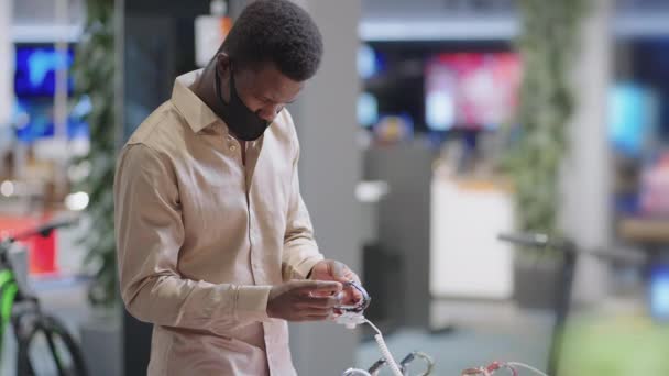 Jovem negro cara está comprando na loja de eletrônicos durante pandemia, vestindo máscara no rosto, escolhendo smartwatch — Vídeo de Stock