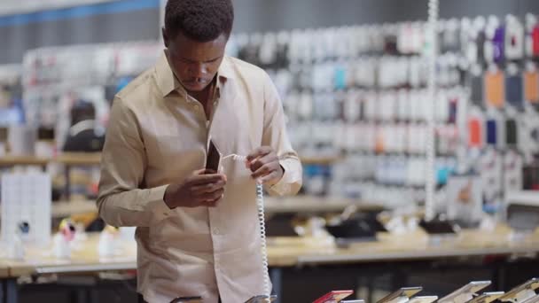 Afro-american guy is shopping in digital equipment store, viewing new models of smartphones in demonstration showcase in trading hall — Stock Video