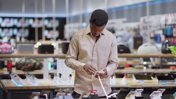 Sklep z cyfrowym sprzętem, mężczyzna kupujący testuje nowy model smartfona w hali handlowej, facet trzyma nowoczesne urządzenie w rękach — Wideo stockowe