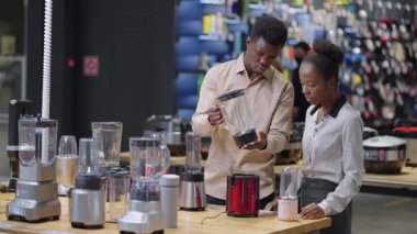 Ev gereçleri mağazasından alışveriş yapanlar, siyah adam ve kadın blender örneklerini inceliyor, evli çift tartışıyor.
