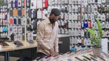 Yüzü maskeli Afro-Amerikalı adam büyük dijital ekipman mağazasından alışveriş yapıyor. Gösteri salonundaki akıllı telefonların yeni modellerine bakıyor.