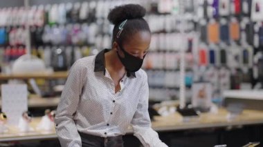 Yüz maskeli genç siyahi bayan dijital ekipman dükkanından akıllı telefon alıyor, kadın ticaret salonunda yeni modeller izliyor.