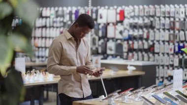 Zenci adam cep telefonu dükkanında alışveriş yapıyor, büyük perakende satış salonundaki modern akıllı telefonu test ediyor.