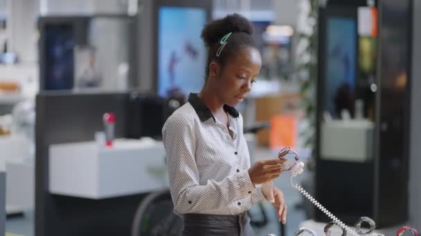 Чорна жінка в магазині цифрового обладнання, жінка-покупець вибирає нову модель смарт-годинника, переглядаючи зразок на демонстраційній вітрині — стокове відео