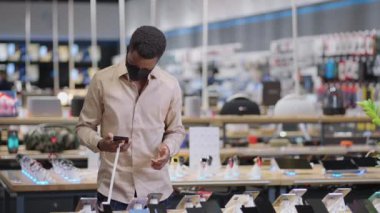 Yüz maskeli siyah adam dijital ekipman mağazasında akıllı telefonu test ediyor. Satın almadan önce yeni modellere bakıyor. Erkek alışverişçinin portresi.