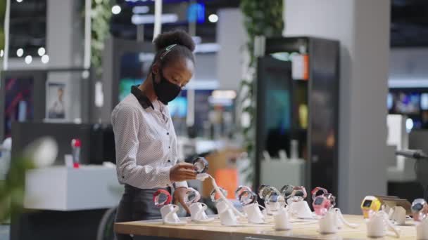 Jovem afro-americana senhora está vendo amostras de exposição de smartwatch na loja de eletrônicos, examinando novos modelos — Vídeo de Stock