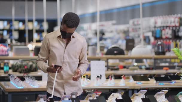 Czarny człowiek w masce testuje smartfona w cyfrowym sklepie sprzętu, oglądanie nowych modeli przed zakupem, portret kupującego — Wideo stockowe