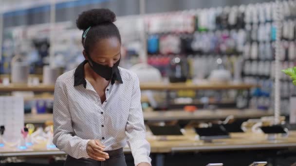 Černá žena s obličejovou maskou testuje smartphone v obchodě s digitálním vybavením, prohlížení nových modelů před zakoupením, portrét ženy nakupující — Stock video