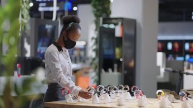 Yüz maskeli siyahi kadın, salgın sırasında elektronik mağazasından akıllı kaset alıyor. Gösteri vitrininde modelleri izliyor.