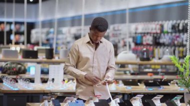Afro-Amerikalı adam dijital ekipman dükkanında akıllı telefonu seçiyor. Ticaret salonundaki erkek alışverişçinin portresi.