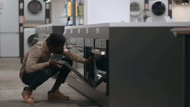 Schwarzer Mann mit Gesichtsmaske sieht Elektroofen in Haushaltsgerätegeschäft, afroamerikanischer Mann sieht Ausstellungsbeispiel in Handelshalle — Stockvideo