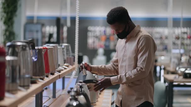 Uomo nero con maschera viso sta esaminando modelli di bollitore elettrico nel negozio di elettrodomestici, l'uomo afro americano sta visualizzando campione mostra sulla vetrina — Video Stock