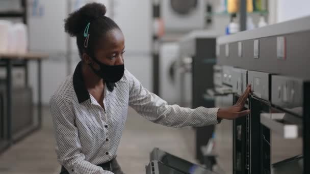 Αφρο αμερικανική γυναίκα με μάσκα προσώπου επιλέγει ηλεκτρικό φούρνο στο κατάστημα συσκευών κουζίνας, θηλυκό αγοραστής είναι προβολή δείγμα έκθεση — Αρχείο Βίντεο