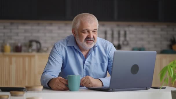 Homme d'âge moyen est bavarder par appel vidéo par ordinateur portable, assis seul à la maison cuisine, boire du thé — Video