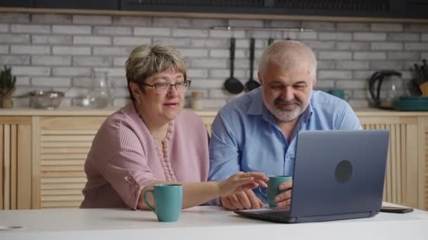 Счастливый пожилой мужчина и женщина в чате онлайн на ноутбуке с друзьями или семьей, портрет на домашней кухне — стоковое видео