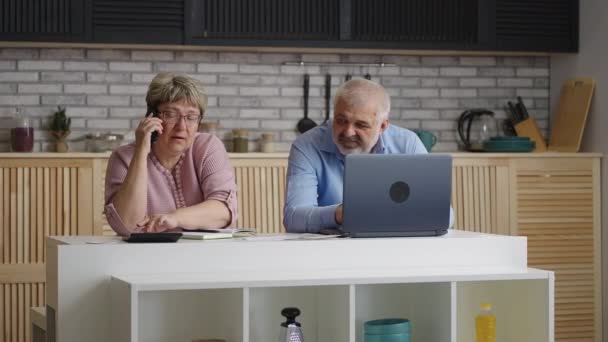 Ama de casa de edad está llamando a la empresa de servicios públicos y la consulta sobre el pago, hombre de edad avanzada está sentado cerca y navegar por Internet por ordenador portátil — Vídeos de Stock