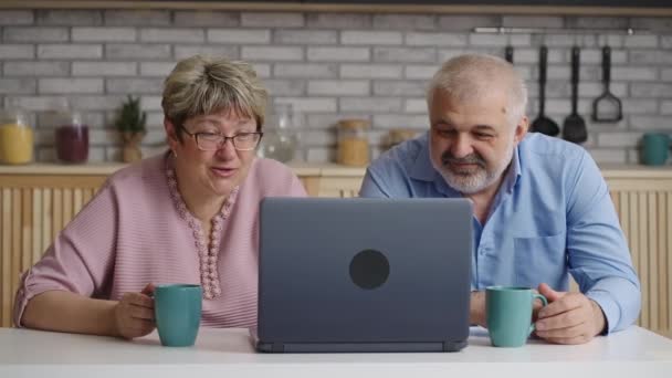 Anciano hombre y mujer están bebiendo té en casa cocina y ver películas o imágenes en la pantalla del ordenador portátil — Vídeos de Stock
