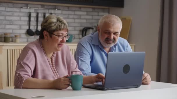 Vieil homme et la femme apprennent à utiliser un ordinateur portable ensemble, assis à la maison cuisine le week-end, en appuyant sur les boutons et en regardant l'affichage — Video