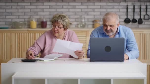 Älteres Ehepaar prüft Stromrechnungen, Frau und Mann betrachten Papier, rechnen mit Taschenrechner und bezahlen online per Laptop — Stockvideo