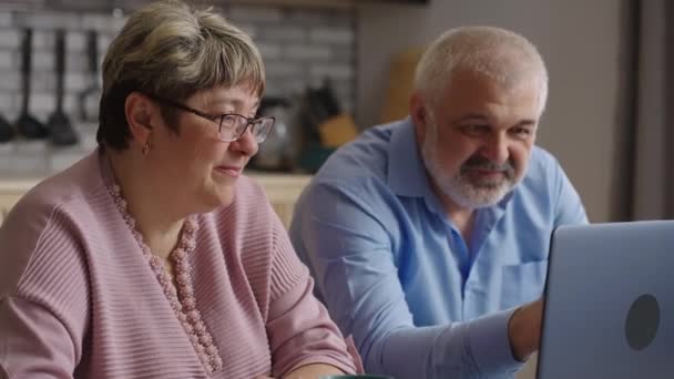 退休人员正在学习使用笔记本电脑，在家里厨房里用现代笔记本电脑上网，笑着的年长配偶正在看屏幕 — 图库视频影像