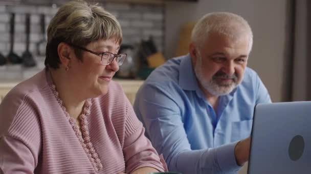 Retraités apprennent à utiliser un ordinateur portable, surfer sur Internet par ordinateur portable moderne à la maison cuisine, souriants conjoints âgés regardent l'écran — Video