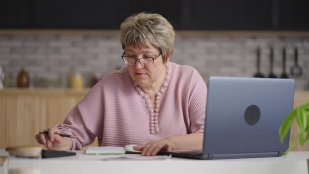 Starsza kobieta oblicza rachunki za media w kuchni domowej, używając kalkulatora i laptopa, sprzątania i finansów — Wideo stockowe