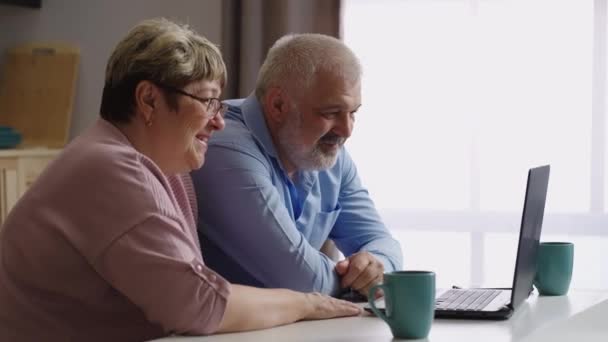Vieil homme et femme communiquent avec des amis en ligne par ordinateur portable, assis à la cuisine, portrait moyen — Video