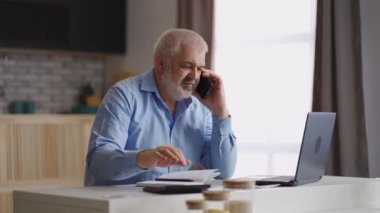 Deneyimli finansal danışman evden çalışıyor, telefonla iletişim kuruyor ve hesaplıyor, hesap makinesi ve dizüstü bilgisayar kullanıyor, fazla mesai yapıyor