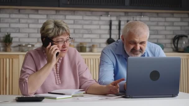 Para emerytów konsultuje się przez telefon, stara kobieta dzwoni i rozmawia, mąż surfuje po Internecie przez laptopa — Wideo stockowe