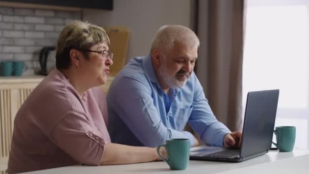 Äldre makar chattar online med vänner eller familj, sitter tillsammans i köket på helgen, välkomnande samtalspartner — Stockvideo